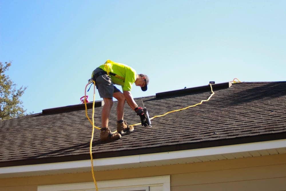 Roofing contractor in a safety harness nailing shingles on a residential roof against a blue sky